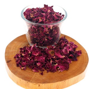 May include: A clear glass container filled with dried deep magenta rose petals, with additional loose dried rose petals scattered around the base of the glass on a circular wooden slice with visible natural wood grain and texture.