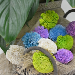Puede incluir: Una colección de flores de sola de madera coloridas en una variedad de tonos, incluyendo blanco, azul claro, morado, verde y verde claro. Las flores están dispuestas en una cesta rústica con tela de arpillera.