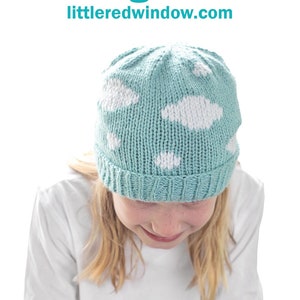 May include: A child wearing a light blue knitted hat with white cloud designs. The hat has a ribbed brim and is made with a soft yarn. The text "Blue Sky Hat Knitting Pattern littleredwindow.com" is visible at the top of the image.