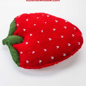May include: A knitted strawberry toy with a green stem and white heart-shaped details. The toy is red and has a soft, squishy texture. The text "Big Squishy Strawberry Knitting Pattern" and "littleredwindow.com" is visible above the toy.