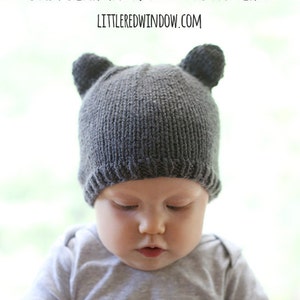 front view of chubby baby in gray short sleeved shirt sitting in a blue chair wearing a dark gray knit hat with bear ears on top and looking down at a camera lens cap in their hands