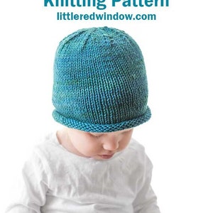 toddler in white shirt and blue pants wearing a knit hat with a rolled brim made of teal blue hand dyed yarn  sitting on a wood table and looking down at their lap