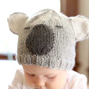 Baby in a pink and white shirt sitting in a blue chair wearing a gray knit hat that looks like a koala looking down and to the left