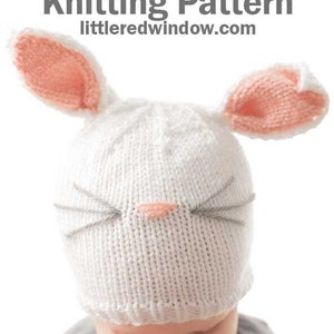 baby in gray shirt wearing a white knit hat with white and pink bunny ears on top and a pink nose and whiskers on the front looking down at their lap