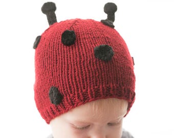 Könnte beinhalten: Eine rote gestrickte Marienkäfermütze mit schwarzen Filzpunkten und Fühler. Die Mütze wird von einem Kind getragen.