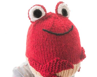 May include: A red knitted crab hat with white eyes and black details. The hat is being worn by a child.