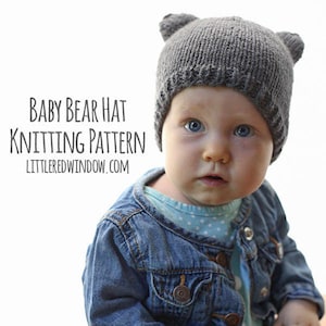 Baby  in a jean jacket and blue onesie holding a lens cap and wearing a dark gray knit hat with bear ears on top sitting on wood in front of a white background