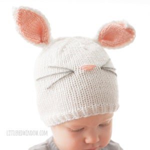 toddler in gray shirt wearing a white knit bunny hat with a pink nose gray whiskers and pink lined bunny ears on top looking down at their hands