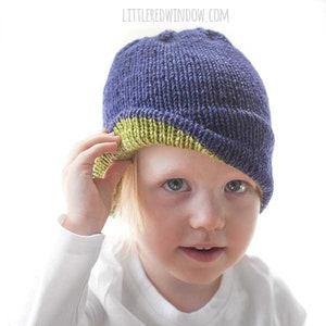 May include: A child wearing a blue and green knitted hat. The hat has a green brim and a blue crown.