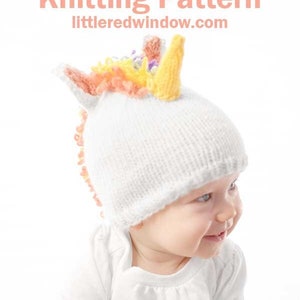 grinning baby in white shirt wearing a white knit hat with golden unicorn horn and rainbow knit mane sitting in front of a white background leaning forward and looking off to the right