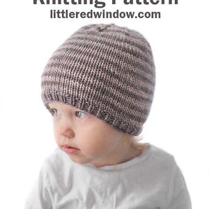 toddler in white shirt wearing a tan and medium brown thin striped knit hat looking to the left in front of a white background