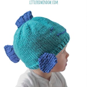 May include: A teal blue knitted hat shaped like a fish with blue fin details. The hat is being worn by a child.