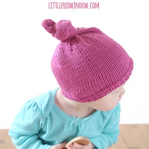 May include: A pink knitted hat with a knot on top. The hat is being worn by a baby sitting on a wooden floor. The text "LITTLE KNIT TOP KNOT HAT KNITTING PATTERN LITTLE RED WINDOW.COM" is visible at the top of the image.