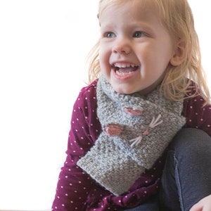 Op de afbeelding: Een jong kind draagt een grijze gebreide sjaal met een kattengezichtontwerp. De sjaal heeft roze oren, een roze neus en witte snorharen. Het kind draagt een bordeauxrood shirt met witte stippen en een blauwe spijkerbroek. Het kind glimlacht en kijkt naar de zijkant.