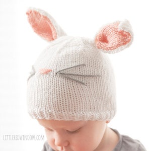 toddler in gray shirt wearing a white knit bunny hat with a pink nose gray whiskers and pink lined bunny ears on top looking down to the left