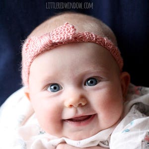 smiling chubby baby with wide blue eyes wearing a light pink ribbed knit headhand with a knit bow on the front being held in front of a dark navy blue background