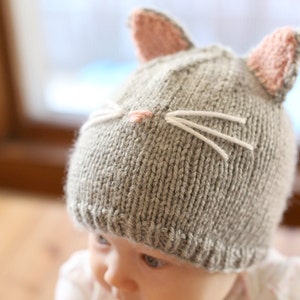 view  of baby in gray onesie wearing a gray knit hat with pink cat ears on top and a little pink nose and white whiskers on the front, looking off to the left while sitting a blue bumbo chair