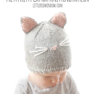 baby in gray onesie wearing a gray knit hat with pink cat ears on top and a little pink nose and white whiskers on the front, looking down