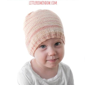 May include: A young child wearing a pink and white striped knitted hat. The hat is made with a soft, textured yarn. The hat is a simple design with a ribbed brim.
