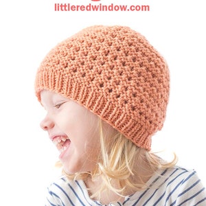 May include: A young child wearing a peach-colored knitted hat with a lace grid pattern. The hat is made with a simple stitch and has a wide brim. The child is smiling and looking to the side. The text "Lace Grid Hat Knitting Pattern" and "littleredwindow.com" is visible at the top of the image.