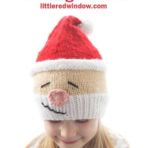 Op de afbeelding: Een kind draagt ​​een gebreide kerstmanmuts met een glimlachend gezicht. De muts is rood en wit met een witte pompon bovenop. Het kind kijkt naar beneden en glimlacht. De muts is verkrijgbaar als breipatroon op littleredwindow.com.