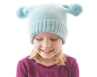 May include: A light blue knitted hat with two fluffy pom-poms on top. The hat is worn by a child with blonde hair and a purple shirt. The text "Double Pom Pom Hat Knitting Pattern littleredwindow.com" is displayed above the hat.