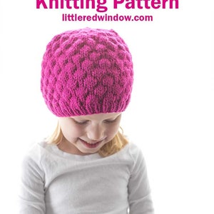 May include: A young person wearing a pink knitted hat with a textured pattern. The hat is called a Raspberry Hat and the knitting pattern is available on the website little red window.com.