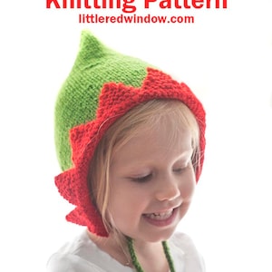 May include: A child wearing a green and red knitted elf pixie bonnet with a pointed top and a scalloped edge. The bonnet has a green tie.