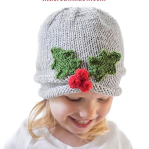 May include: A gray knitted hat with green holly leaves and a red berry applique. The hat is worn by a child with blonde hair. The text "Jolly Holly Hat Knitting Pattern" and "littleredwindow.com" is displayed above the hat.