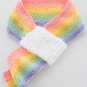 May include: A rainbow colored knitted scarf with a white fuzzy trim. The scarf is folded in half and tied in a knot. The text "Rainbow Scarf Knitting Pattern littleredwindow.com" is at the top of the image.