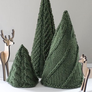 Three dark green knit cone shaped knit Christmas Trees with three different cable knit stitch patterns on a white table with two small wooden deer in front of a gray wall