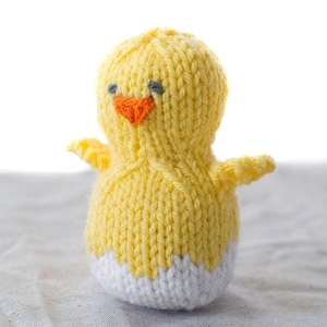 little yellow knit chick with white egg shell pattern on this bottom and two little wings on the sides sitting on a gray table