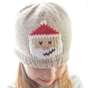 Puede incluir: Un niño o niña lleva una gorra de punto beige con una cara de Papá Noel tejida en la parte delantera. La gorra tiene un gorro rojo, una barba blanca y ojos negros.