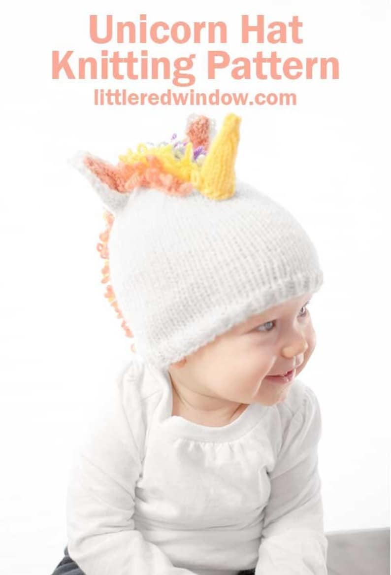 grinning baby in white shirt wearing a white knit hat with golden unicorn horn and rainbow knit mane sitting in front of a white background leaning forward and looking off to the right