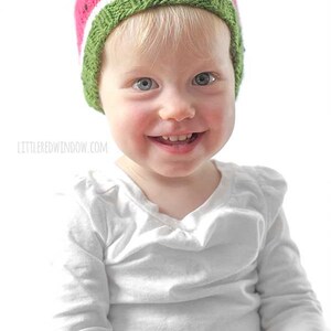 smiling baby in white shirt wearing a knit watermelon hat in green white and pink with black knit seeds in front of a white background