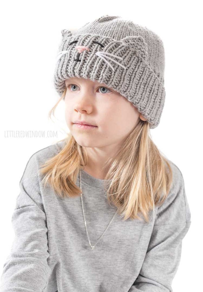 May include: A gray knitted beanie with a cat face design. The cat face has black eyes, a pink nose, and white whiskers.