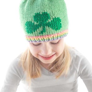 A young girl is wearing a green knit hat featuring a charming shamrock knitting pattern. She is smiling and dressed in a white long-sleeve shirt. The plain white background emphasizes her as the focal point of the image.