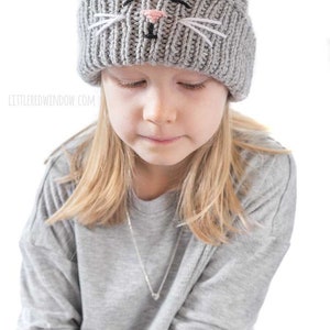 May include: A young person wearing a gray knitted hat with a cat face embroidered on the front. The hat has white whiskers and a pink nose.
