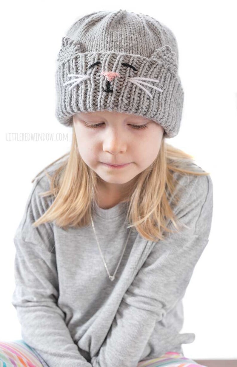 May include: A young person wearing a gray knitted hat with a cat face embroidered on the front. The hat has white whiskers and a pink nose.