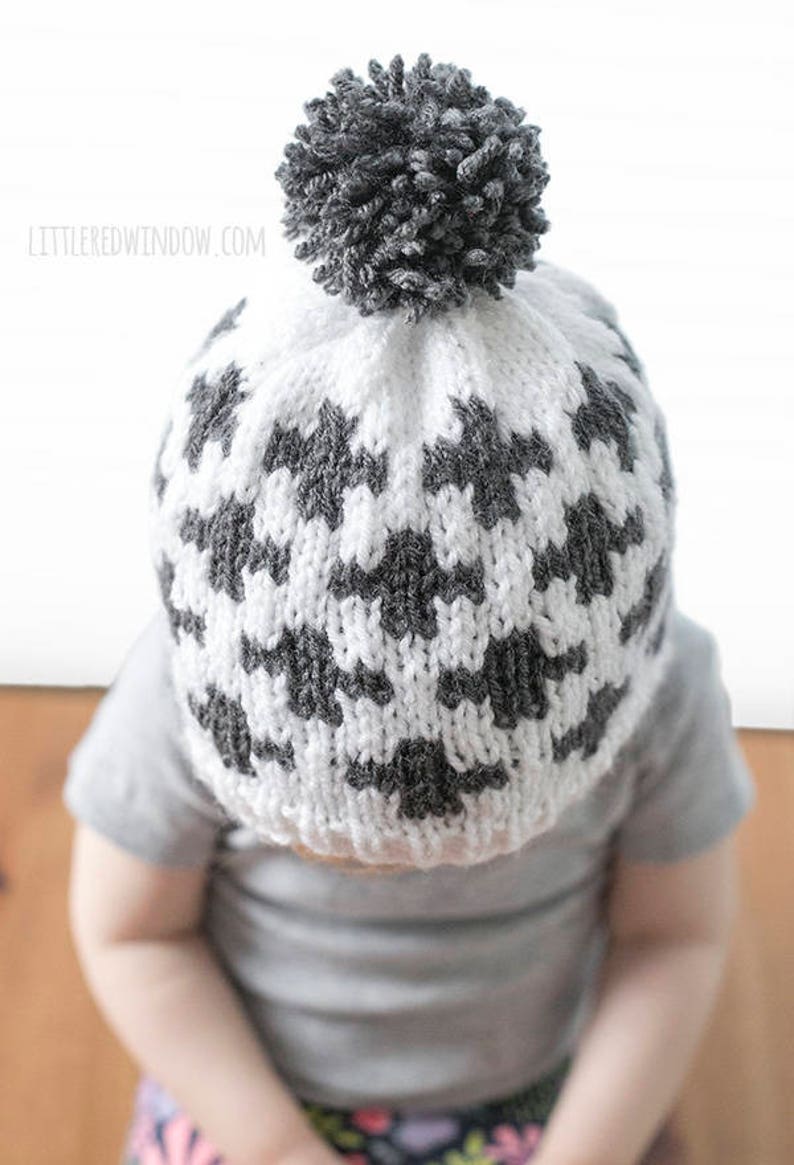 Puede incluir: Un gorro de punto blanco y gris con un pomp&oacute;n gris en la parte superior. El gorro tiene un patr&oacute;n geom&eacute;trico.