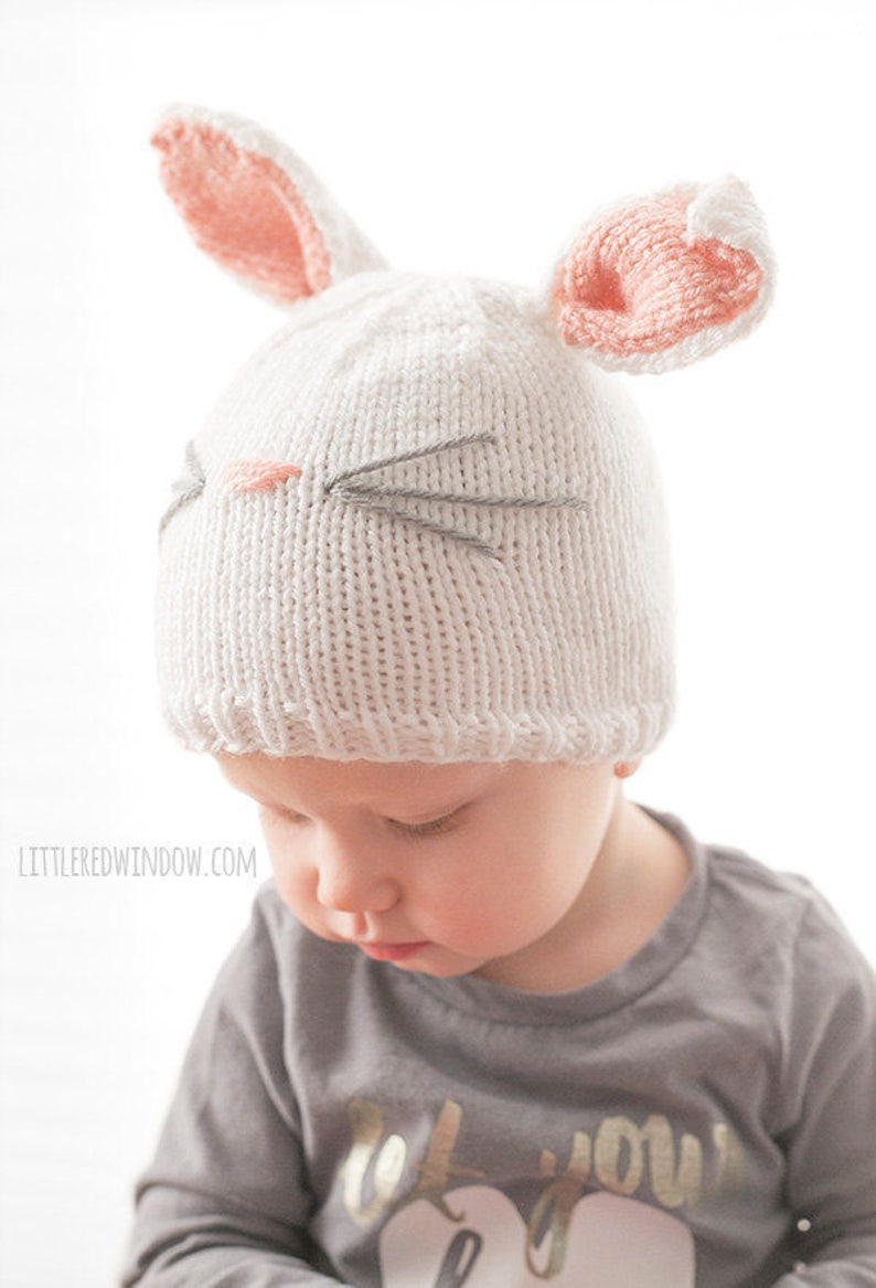 toddler in gray shirt wearing a white knit bunny hat with a pink nose gray whiskers and pink lined bunny ears on top looking down to the left