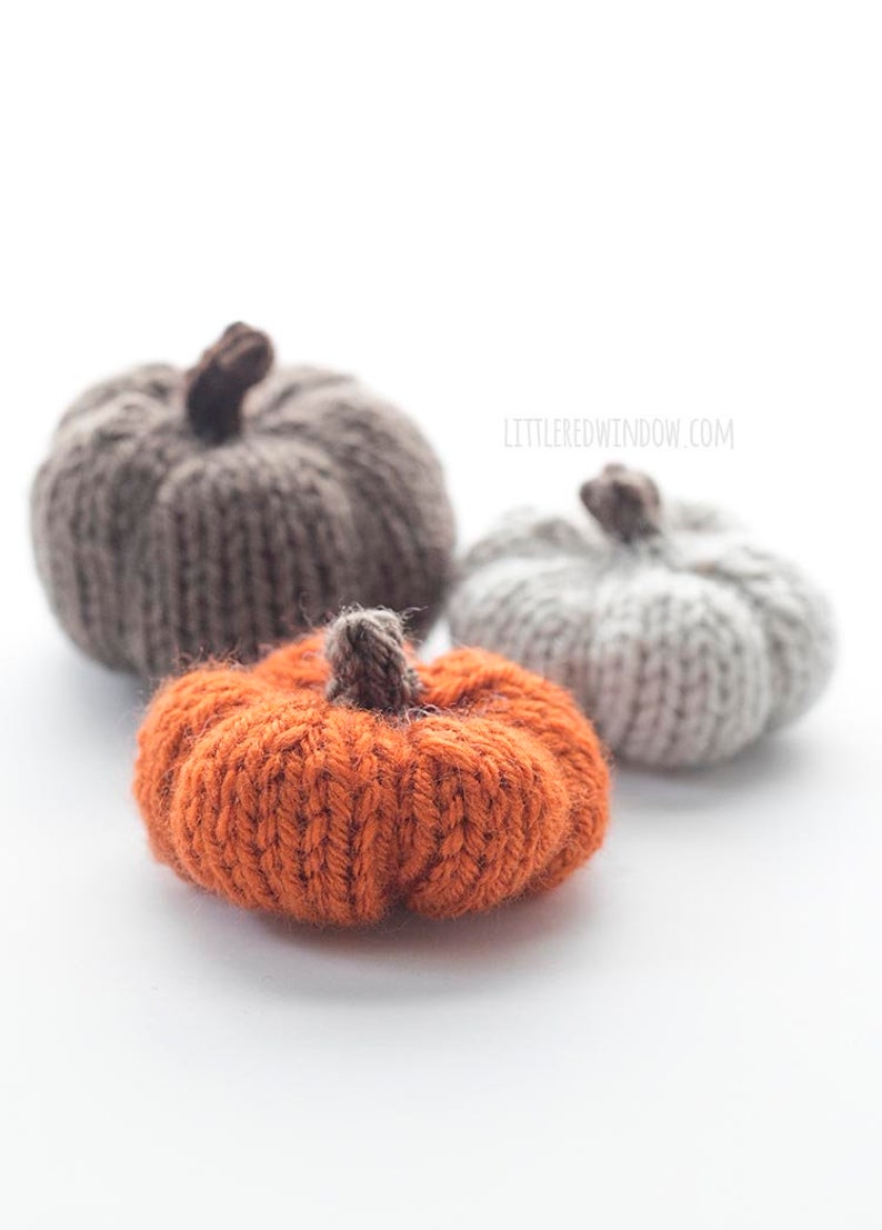 three small knit pumpkins knit with orange tan and medium brown yarn with dark brown knit pumpkin stems sitting on a white table with the orange pumpkin in front