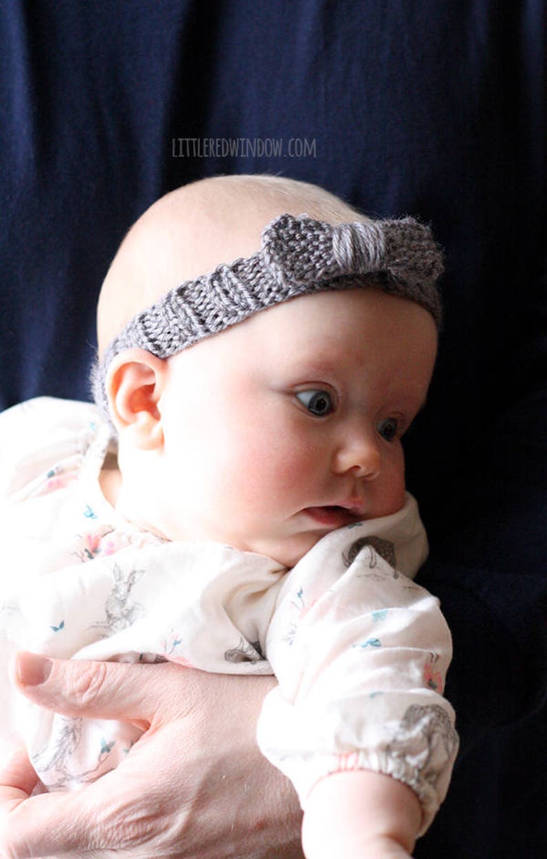 chubby baby with wide blue eyes wearing a medium dusty purple ribbed knit headhand with a purple knit bow on the front and being held in front of a dark navy blue background and looking off to the right
