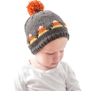 An infant wearing a grey knitted Candy Corn Hat featuring a candy corn pattern and a large orange pom-pom on top. The infant is dressed in a white shirt and looking downward.