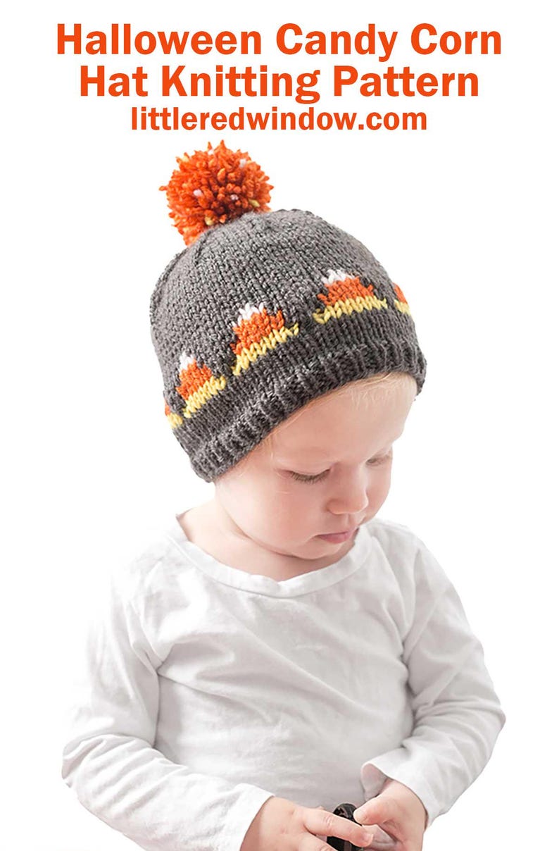An infant wearing a grey knitted Candy Corn Hat featuring a candy corn pattern and a large orange pom-pom on top. The infant is dressed in a white shirt and looking downward.