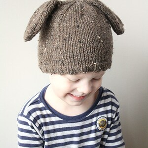 Puede incluir: Un ni&ntilde;o lleva un gorro de punto marr&oacute;n con orejas ca&iacute;das en forma de cachorro. El gorro tiene un patr&oacute;n moteado. El ni&ntilde;o lleva una camisa a rayas azul y blanca.