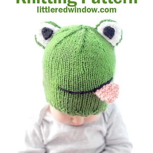 May include: A green knitted frog hat with black eyes, a white tongue, and white accents around the eyes. The hat is being worn by a baby.