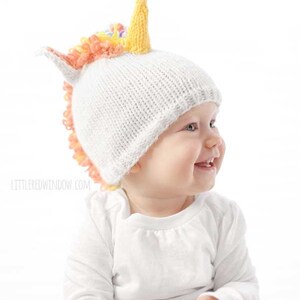 grinning baby in white shirt wearing a white knit hat with golden unicorn horn and rainbow knit mane sitting in front of a white background and looking off to the right