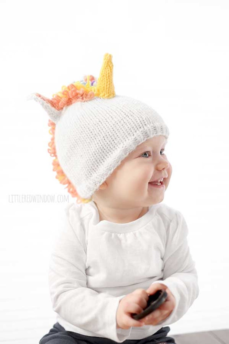 grinning baby in white shirt wearing a white knit hat with golden unicorn horn and rainbow knit mane sitting in front of a white background and looking off to the right