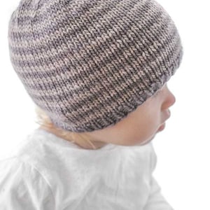 toddler in white shirt wearing a tan and medium brown thin striped knit hat looking off to the right in front of a white background